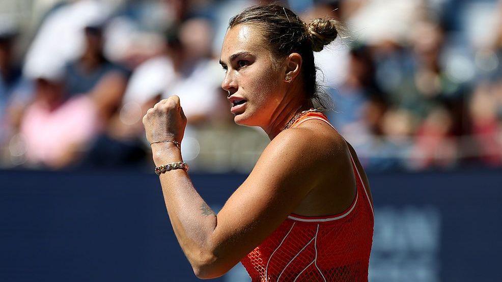 Aryna Sabalenka, wearing an orange sleeveless dress, stands side on and raises a clenched left fist