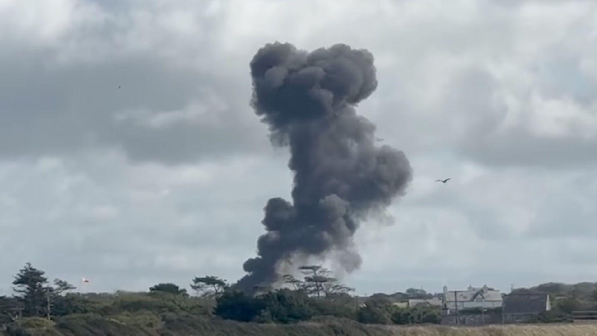 Smoke billows into the sky from the trees and shrubbery, something appears to be on fire in the left hand side. Trees and shrubbery and a house is visible in the foreground.