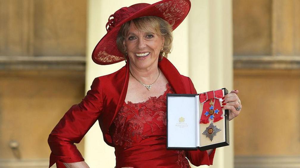 Esther Rantzen poses after she was made a Dame at an investiture ceremony at Buckingham Palace in 2015.  She is holding up her medal to camera and smiling - wearing a red outfit and matching wide brimmed hat.