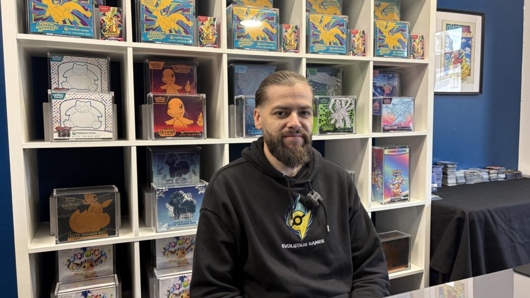 Pokémon fans prepare for ‘biggest day in a decade’ A man with dark hair tied back and a long, dark beard, dressed in a black hoodie smiles at the camera. He is sitting in front of shelving blocks, all of which contain a sealed casing of Pokémon cards or merchandise.
