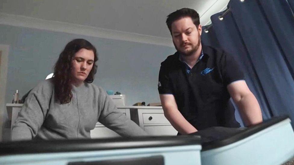 Catie and Phil stand in a bedroom looking down at their open suitcase.