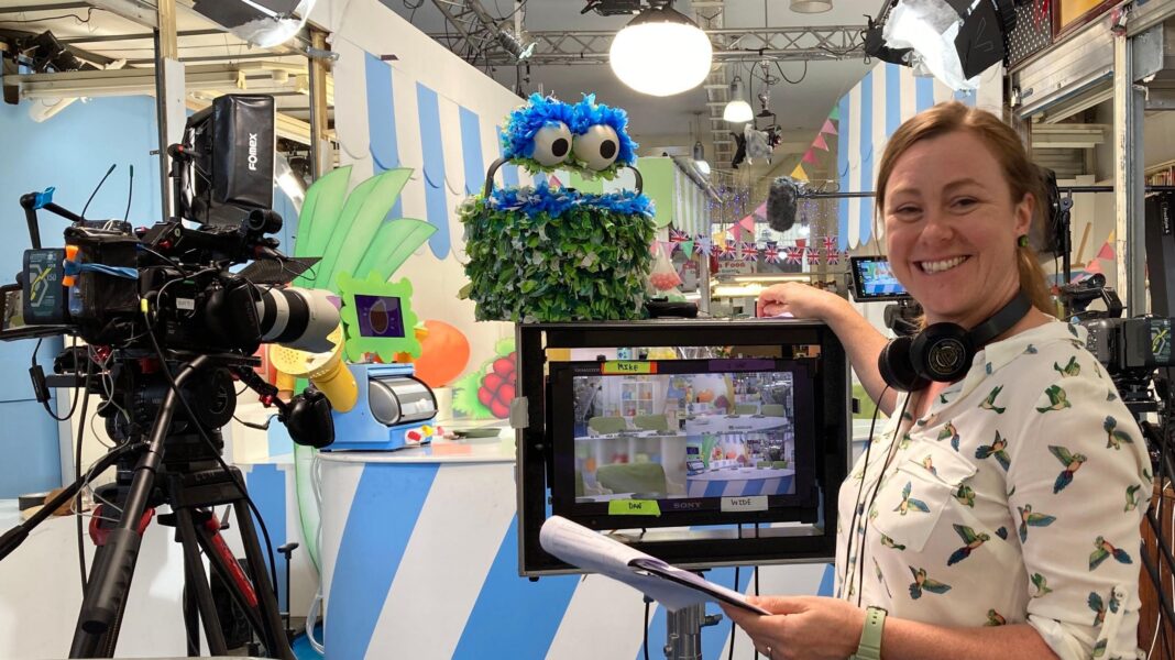 A person wearing headphones around their neck stands next to a monitor and television cameras inside a market-based studio set. The scene includes a green puppet character made from leaf-like material with large blue eyes. Overhead studio lights, microphones and camera equipment are visible, while the monitor shows multiple camera views of the children's programme set. Blue-and-white striped market stall backdrops, fruit-themed decorations and bunting can be seen in the background.
