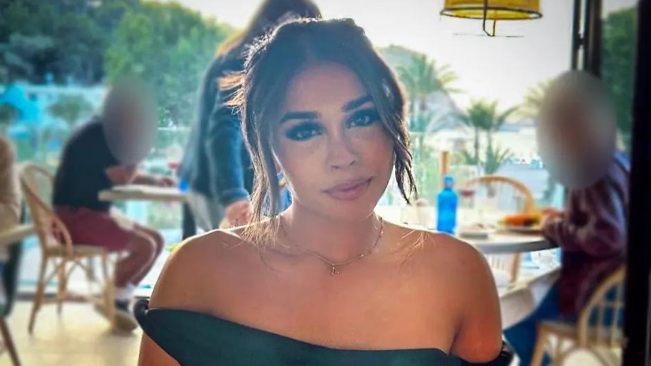 Ravika sitting in a restaurant, pictured head and shoulders looking directly at the camera. She has her hair tied up and is wearing a black off-the-shoulder dress.