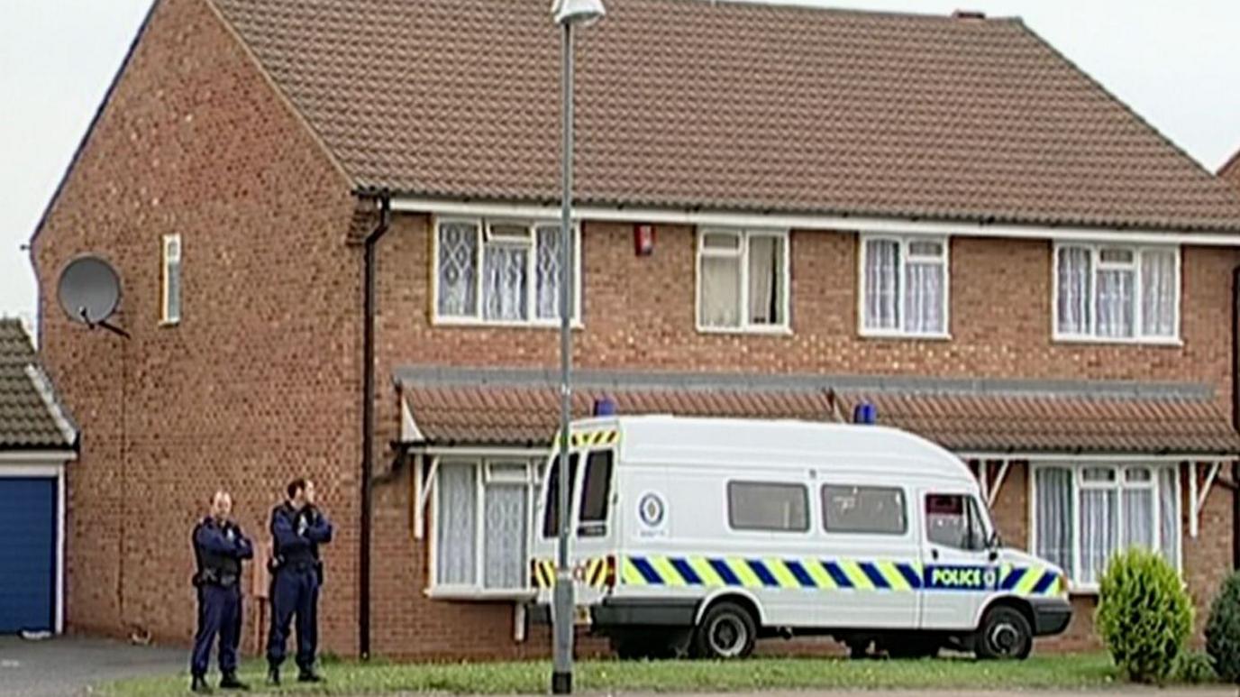 A still from a video showing a small, mid-century semi-detached home in Birmingham with net curtains and a satellite dish. A police van is parked outside, blocking the front door, and two police officers stand to the side of the building, with their arms folded