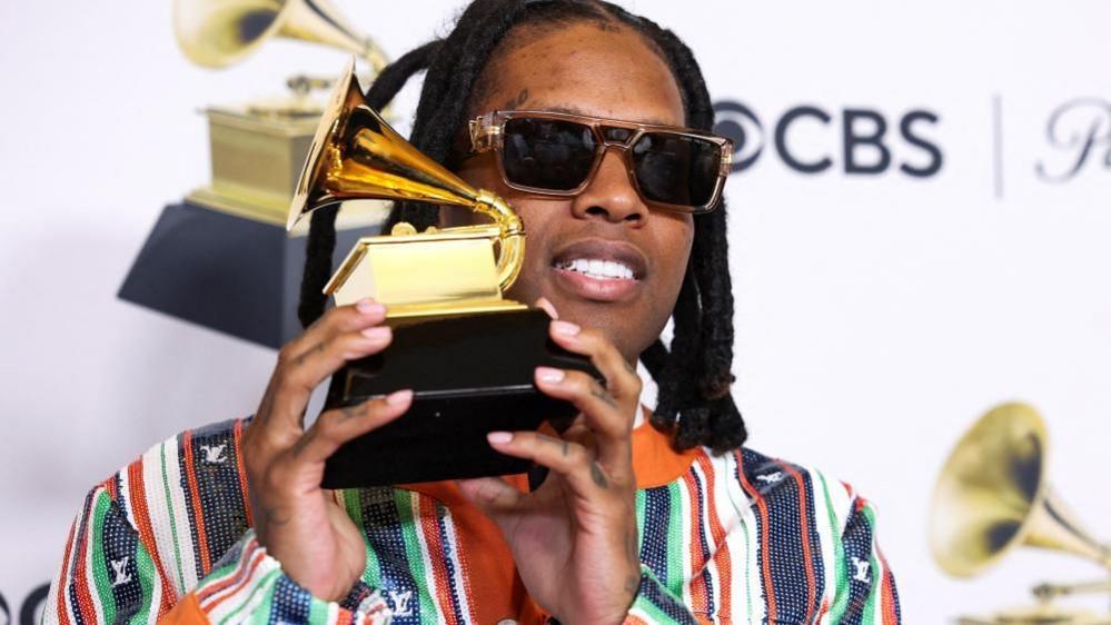 Lil Durk wearing sunglasses and a multi-coloured suit, and holding up the gold gramophone-shaped Grammy Award