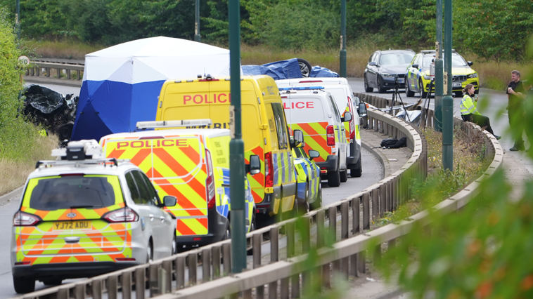 Police vans of varying size are lined up in a row, leading up to the site of the wreckage, all with reflective liveries. To one side, two police officers rest holding paper cups, with one sitting on the central reservation metal crash barrier which separates the two sides of the road.