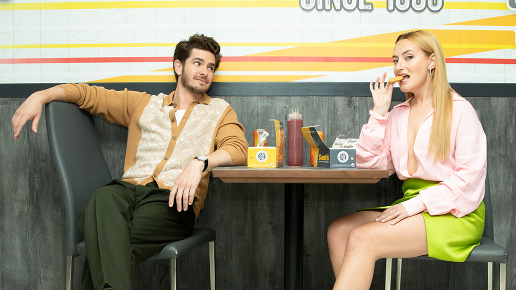 Andrew Garfield sits opposite Dimoldenberg in a chicken shop, looking at her while she bites a chicken nugget. The actor has short curly brown hair and wears a patterned camel cardigan over green trousers. Dimoldenverg wears a low cut pink blouse and neon green skirt.