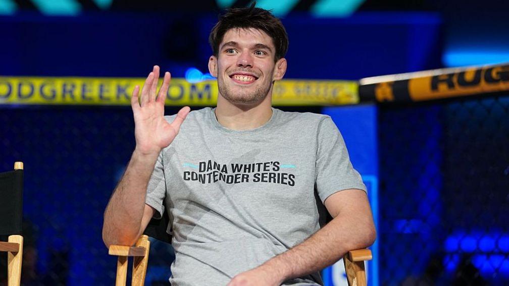 George Staines is smiling and waving as he wears a grey t-shirt