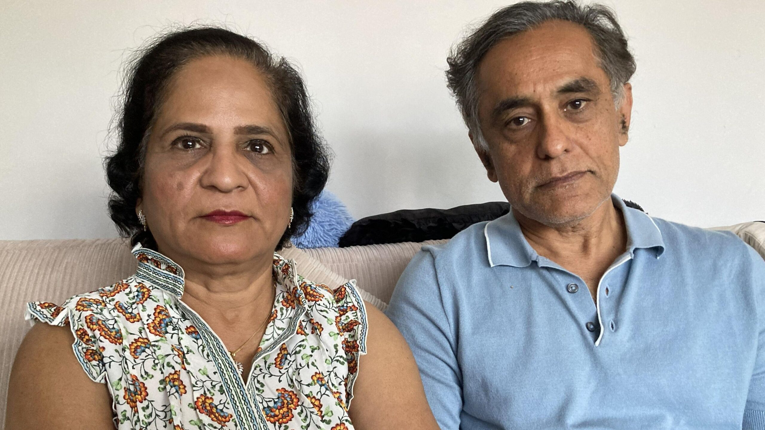 Sandy and Uttam Shiralkar sit on a beige sofa looking at the camera. She has medium-length curly black hair and is wearing a floral patterned top, while he is wearing a light blue polo shirt