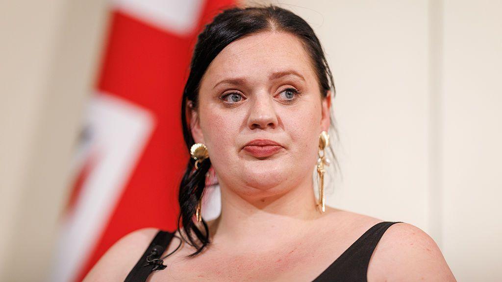 Fiona Goddard, pictured at a press conference in June 2025, has her black hair tied up, wears gold earrings and a black vest top