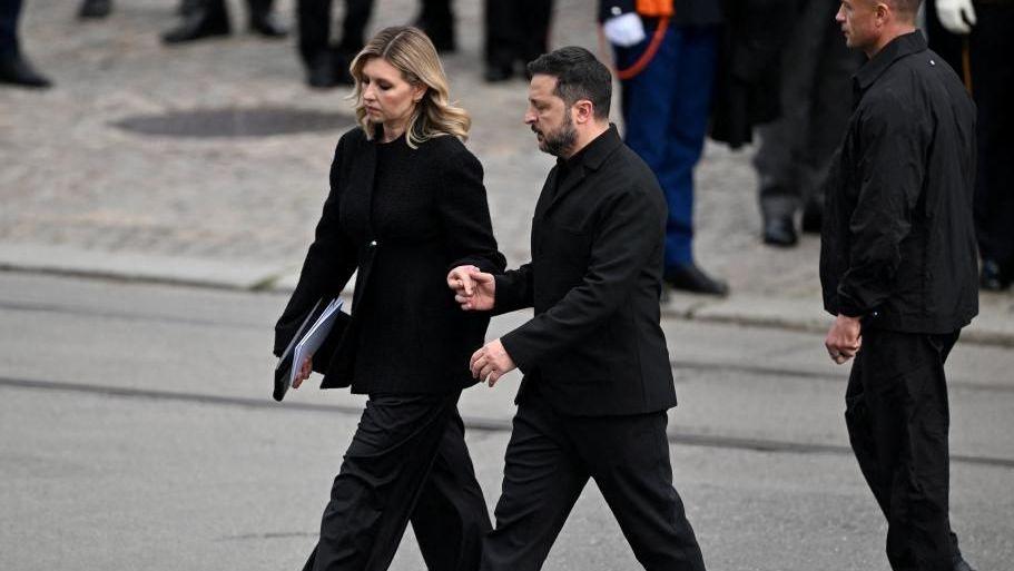 Olena and Volodymyr Zelensky hold hands as they leave the funeral. They are both dressed in black and are outside.