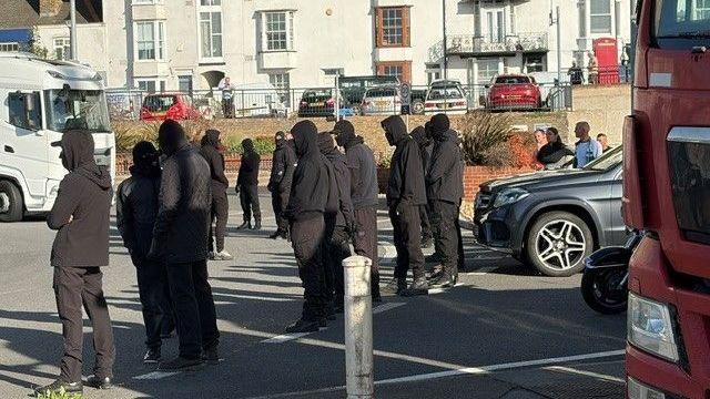 A row of masked men block a roundabout. Cars and lorries are stationary on the road.