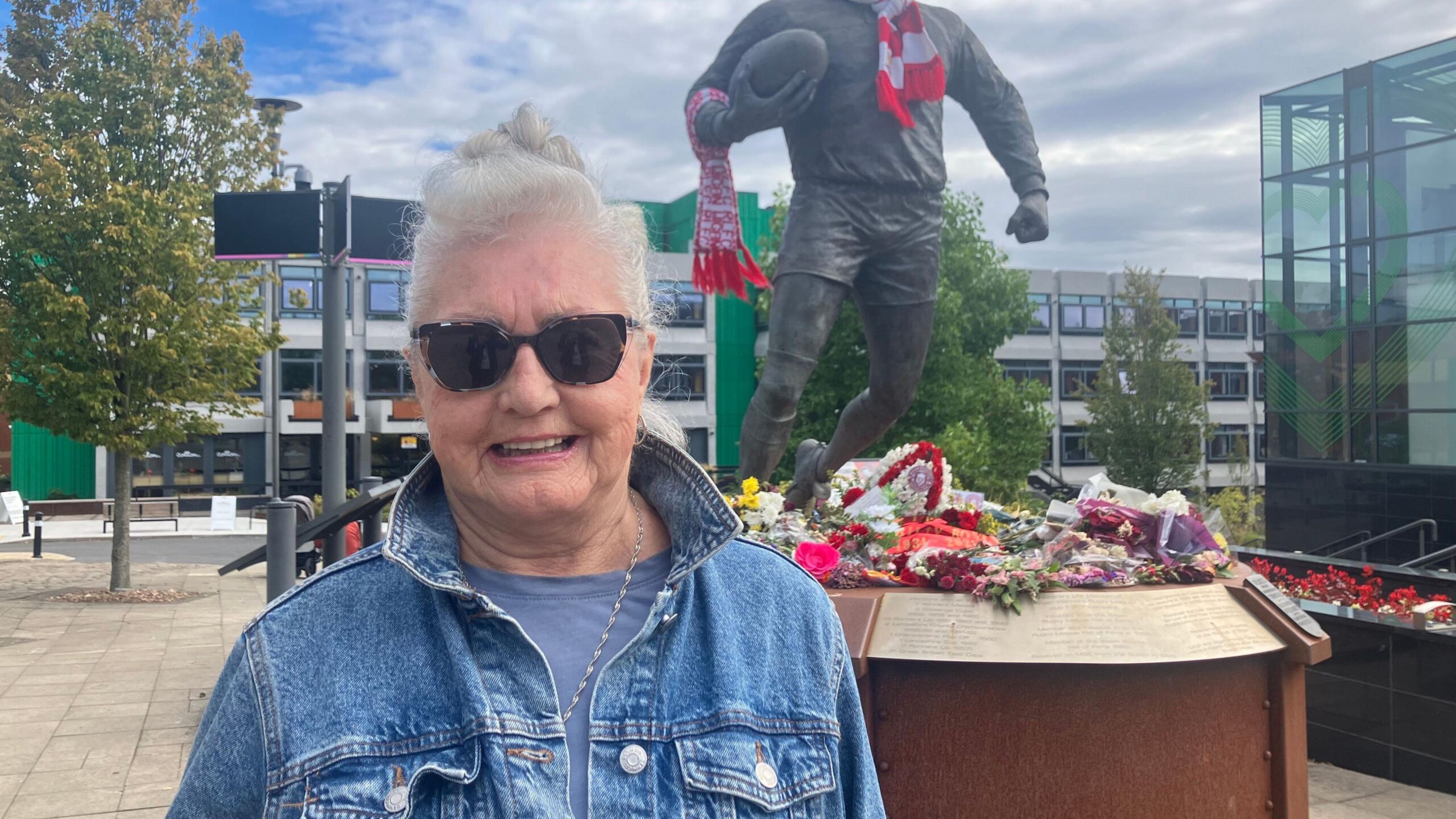 A lady with white hair and sunglasses smiles to the camera. She is wearing a blue denim jacket. Behind her is a statue of Billy Boston holding a rugby ball. He had a red and white scarf around his neck. Flowers surround the base.