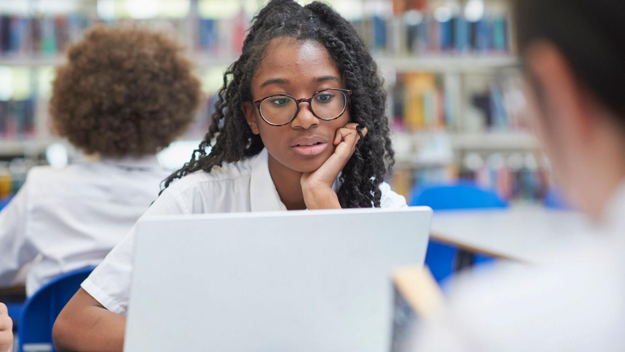 A student wearing glasses uses a laptop in a school library, seated at a table with bookshelves blurred in the background. Other students are visible out of focus nearby.