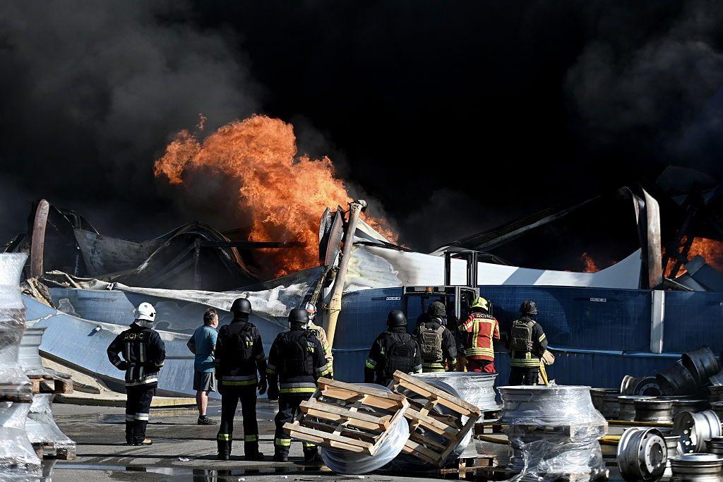 Firefighters watch a blaze amidst wreckage and black smoke.