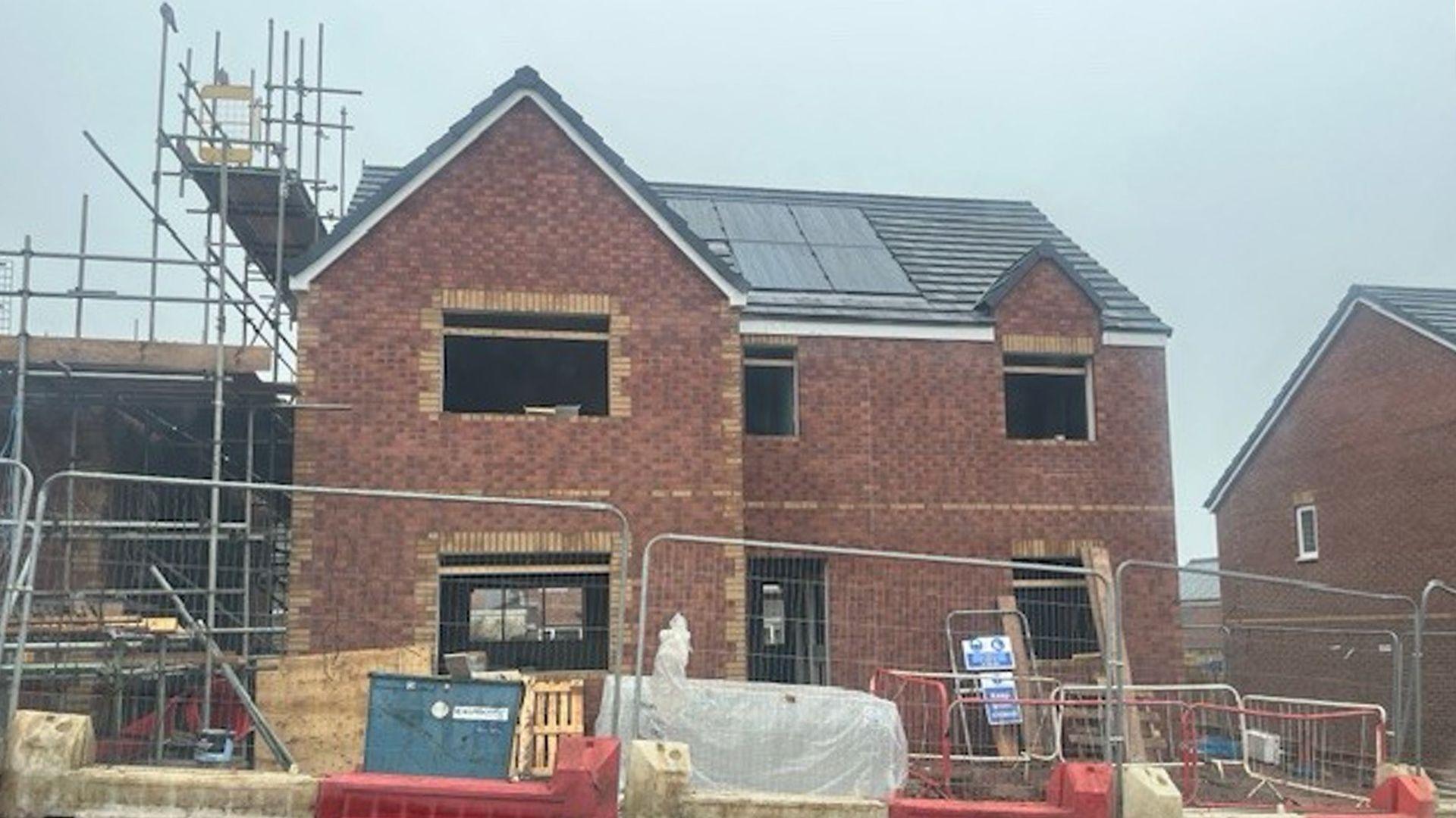 A two‑storey brick house under construction with dark roof tiles and solar panels. Open window and door spaces show the interior unfinished. Scaffolding stands on the left side, while pallets, barriers and wrapped materials sit behind metal fencing at the front.