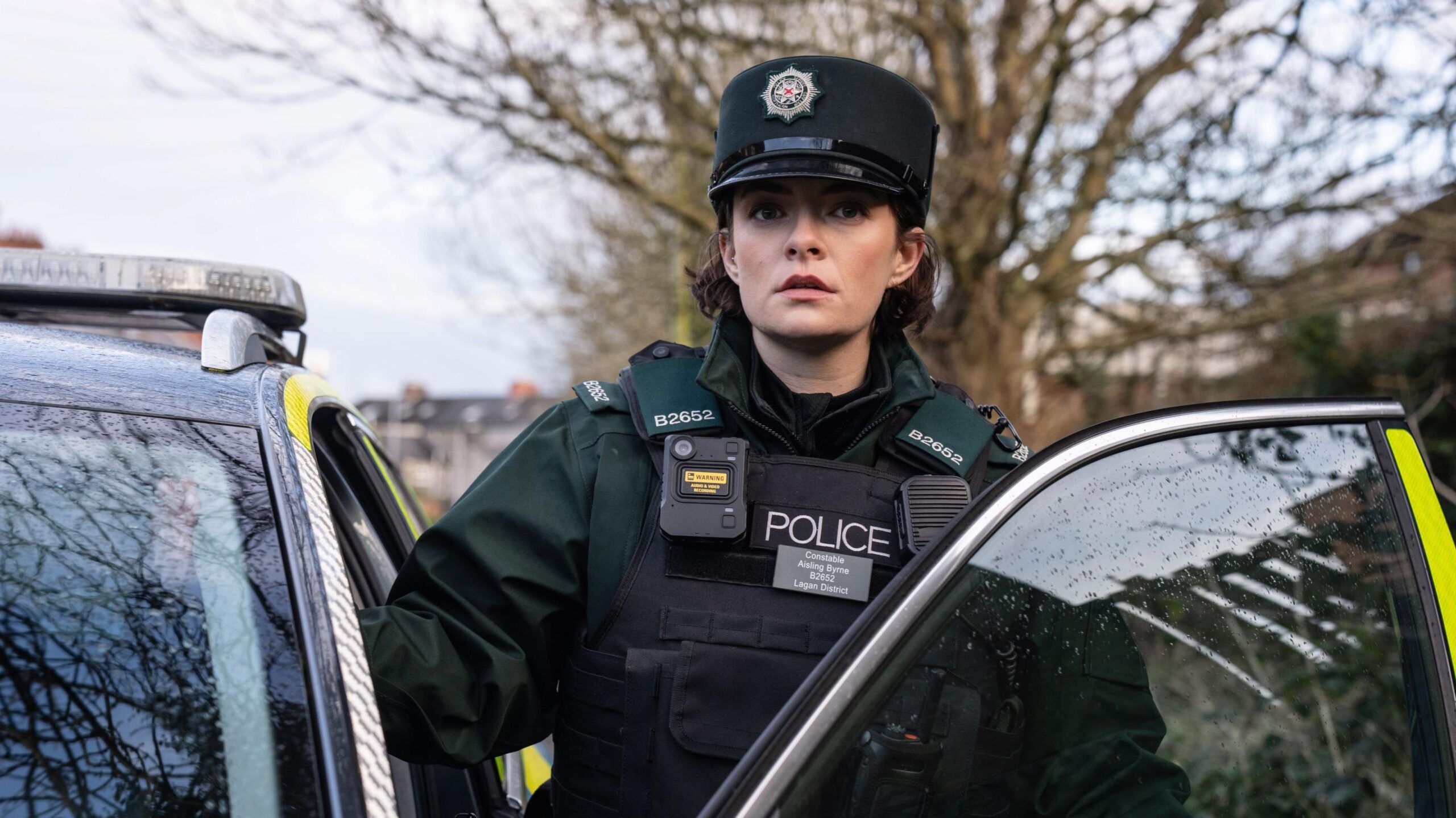 Aisling is wearing a dark green police uniform including a body-cam, stab vest, walkie-talkie and a hat. She is stepping out of the passenger side of a police car. She has dark brown curly hair and is looking straight ahead.