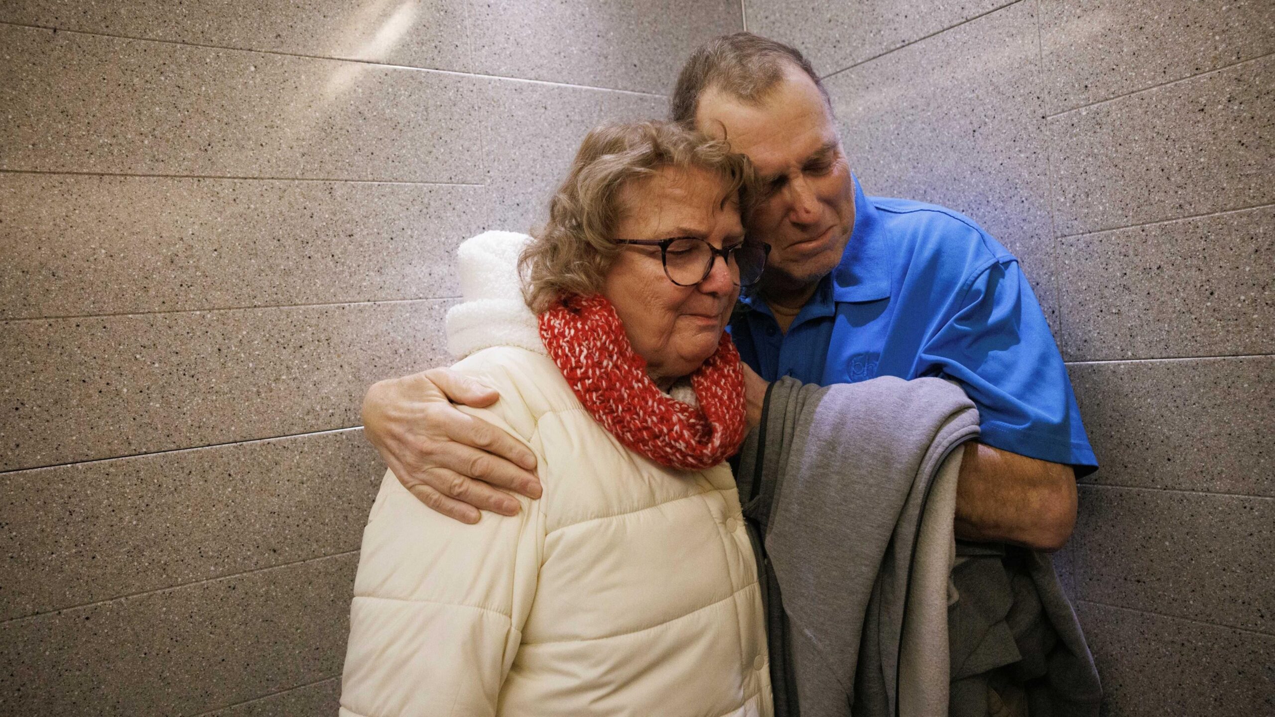 The image shows two adults standing together in a narrow area with grey stone or ceramic tiled walls. One person is wearing a bright blue polo shirt and holding a folded grey garment, while the other is dressed in a light-coloured padded winter coat with a thick red knitted scarf and glasses. The two individuals are positioned very close to one another, with an arm wrapped around the other's shoulder. The composition is tightly framed around the pair, drawing attention to their physical closeness.
