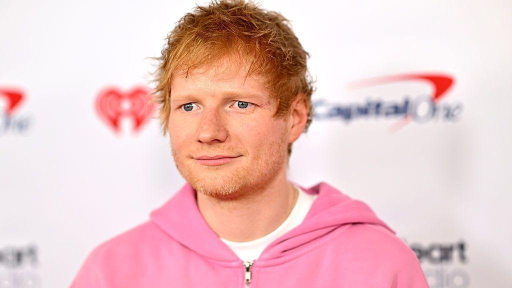 Singer Ed Sheeran poses for red-carpet arrivals in New York in 2025 wearing a pink hoodie and white shirt.