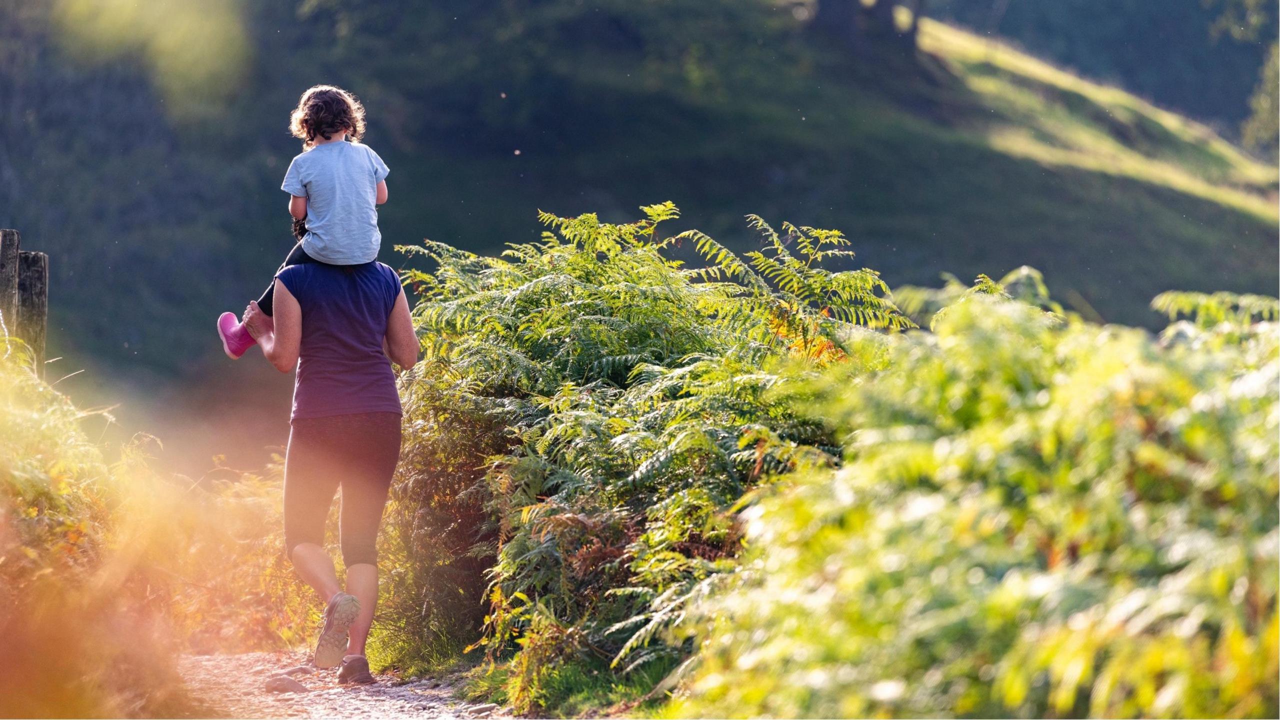Woman carries child, wearing pink wellington boots, on her shoulders past ferns in the sunshine.