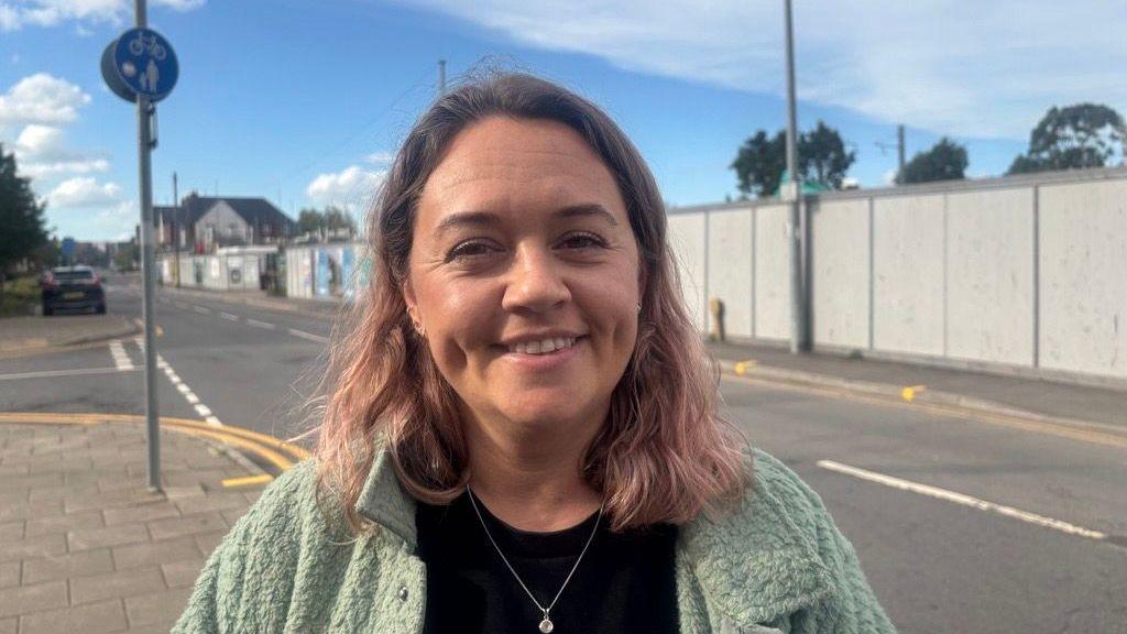 Rhiannon Simmonds stands on the street. She has shoulder length brown hair, wears a black t-shirt and a sage green jacket. Blue sky can be seen behind her.