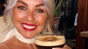 A woman smiles at the camera, she is raising a cocktail glass filled with espresso martini to the camera in a cheers. She has blonde hair and is wearing a white top and neck scarf. She has red lipstick on.