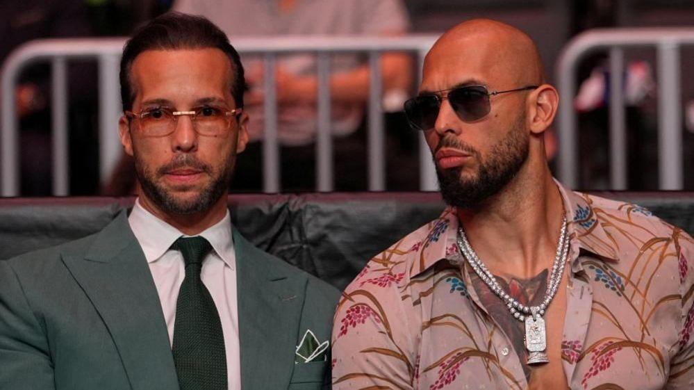 Tristan Tate is wearing sunglasses and is dressed in a green suit and tie and white shirt. Andrew is to his right in sunglasses and an open necked shirt.