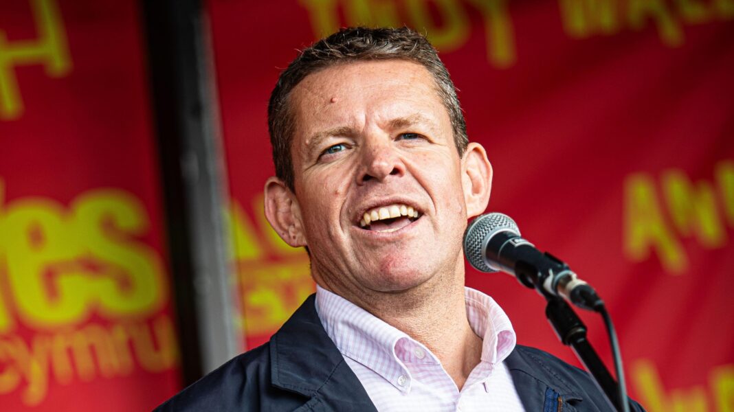 Rhun ap Iorwerth, who has short grey hair and is wearing a salmon pink and white check short and navy blue jacket, speaking into a microphone with red and yello 