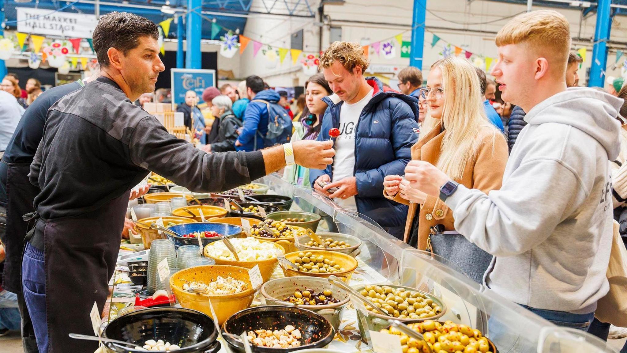 A photo of a bustling food hall with colourful bunting. On the left of the shot a trader stands behind his table which has a range of large ceramic bowls with a range of olives in them. He is reaching across the table to offer a sample of his food to a young man in a grey hoodie. Another three or four people are looking at the produce.