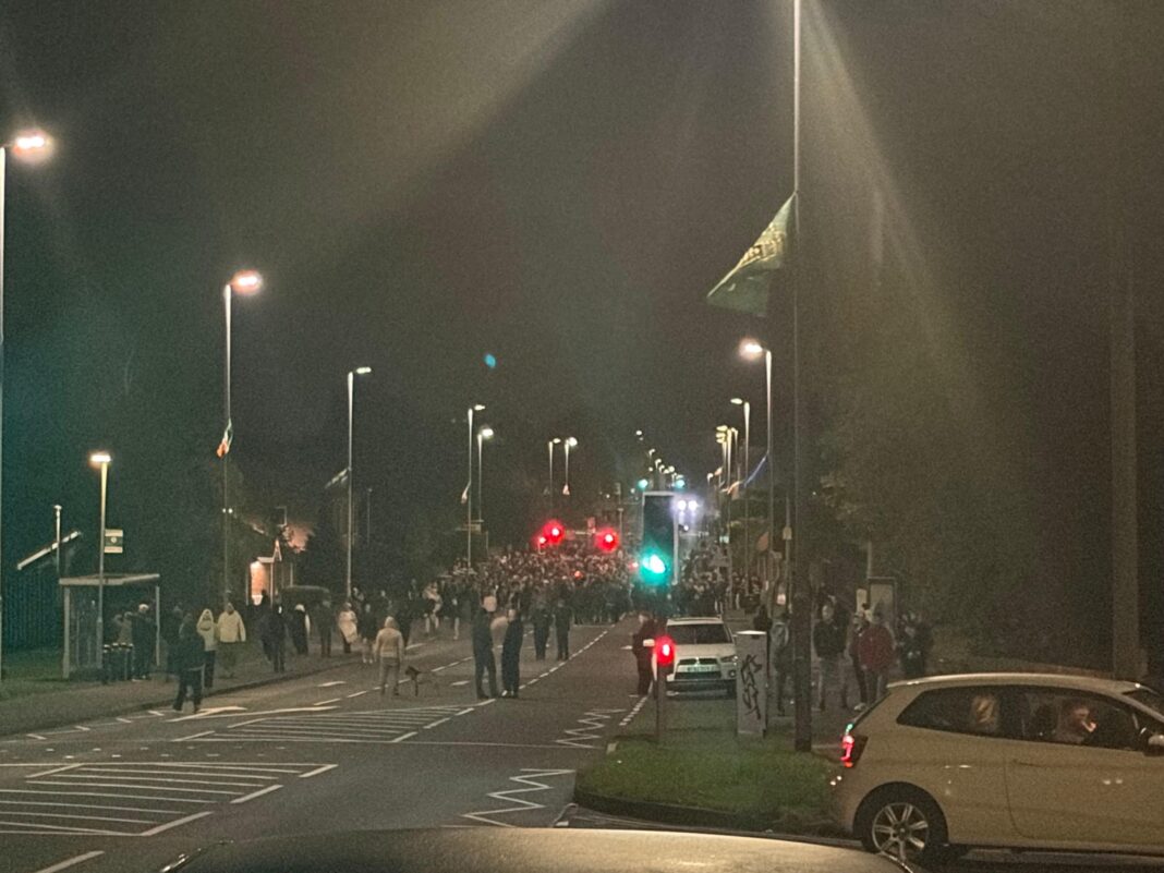 A main road featuring traffic lights and street lights with a large crowd gathered on it in Portadown