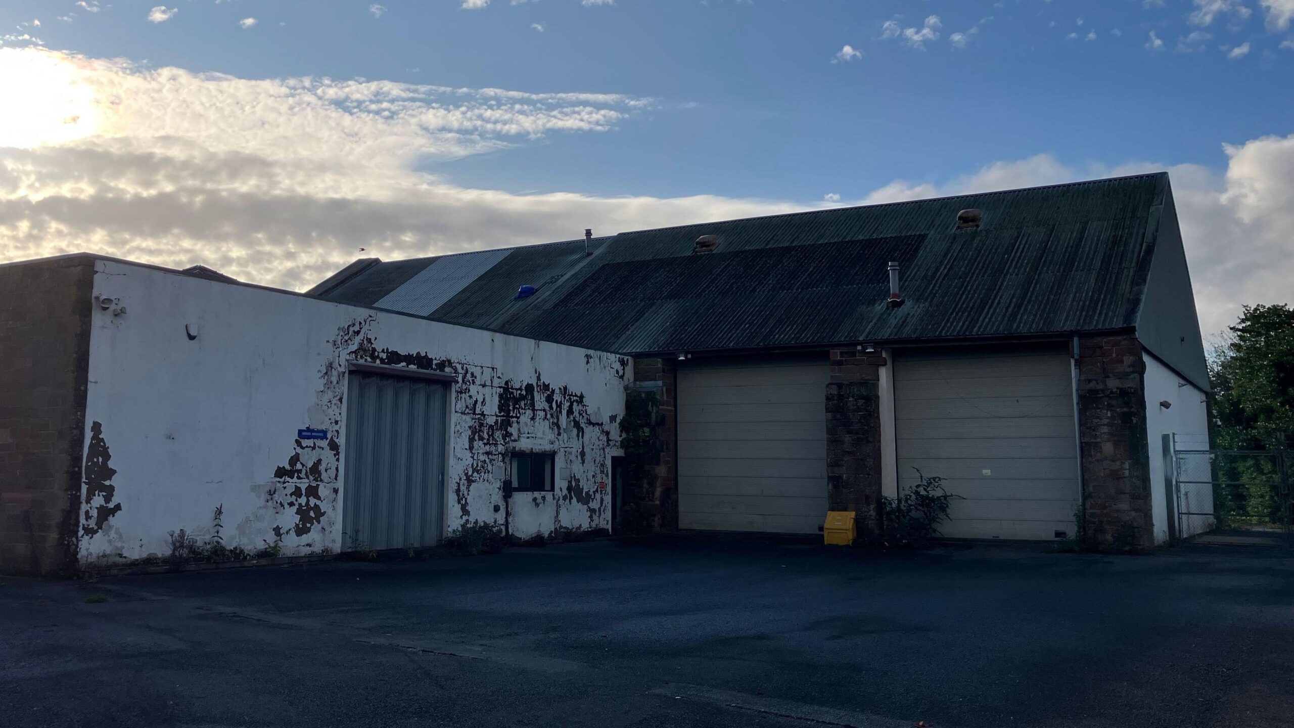 A weathered industrial building with white-painted walls showing extensive peeling paint and exposed masonry. The structure has a large corrugated metal roof, two grey roller-shutter garage doors, and a metal access door. The foreground is a wet asphalt yard, suggesting recent rain. Bright sunlight breaks through clouds on the left, creating strong contrast between the illuminated sky and the darker building, with trees visible at the edge of the site.