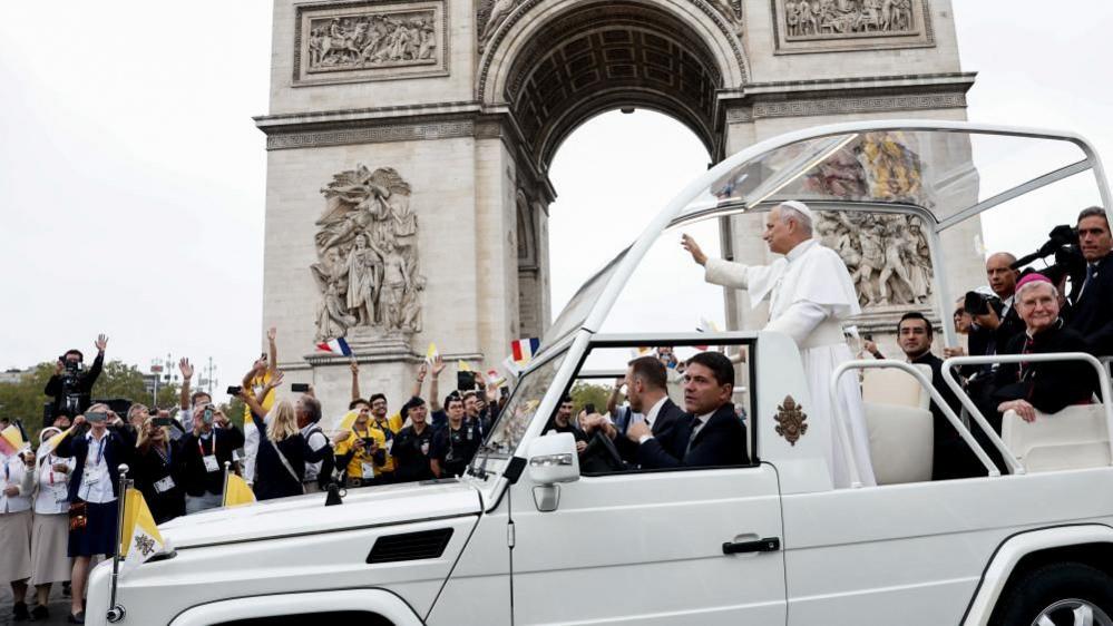 The pope holds his arm up and waves as his popemobile moves past the Arc de Triomphe. The crowd around him waves and hold up French flags