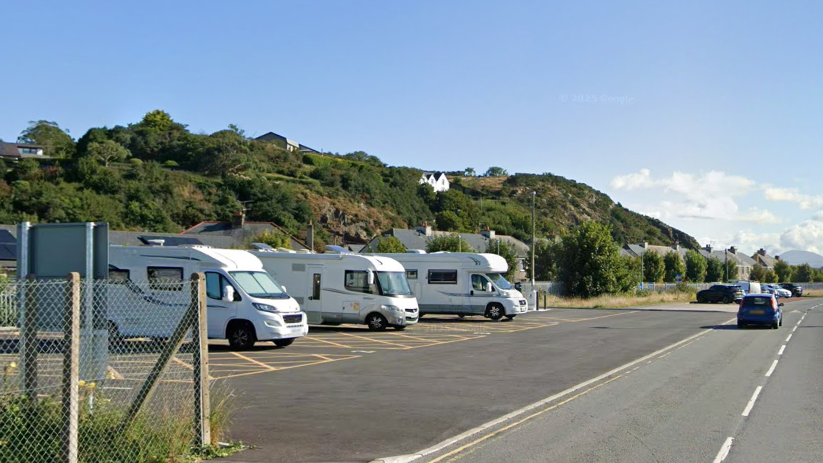 Three motorhomes are parked up in designated bays just over the road from the sea. A car is driving down the road into the distance and some cars can be seen parked in a bay further down the road.