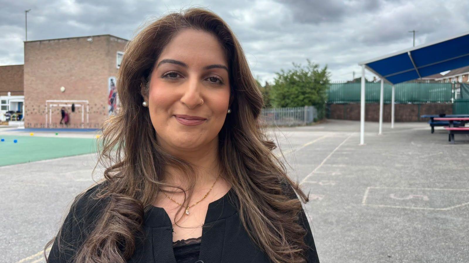 Farzana has long dark hair wearing a black top and necklace, standing in a school playground with a brick building, covered seating area, and cloudy sky in the background.