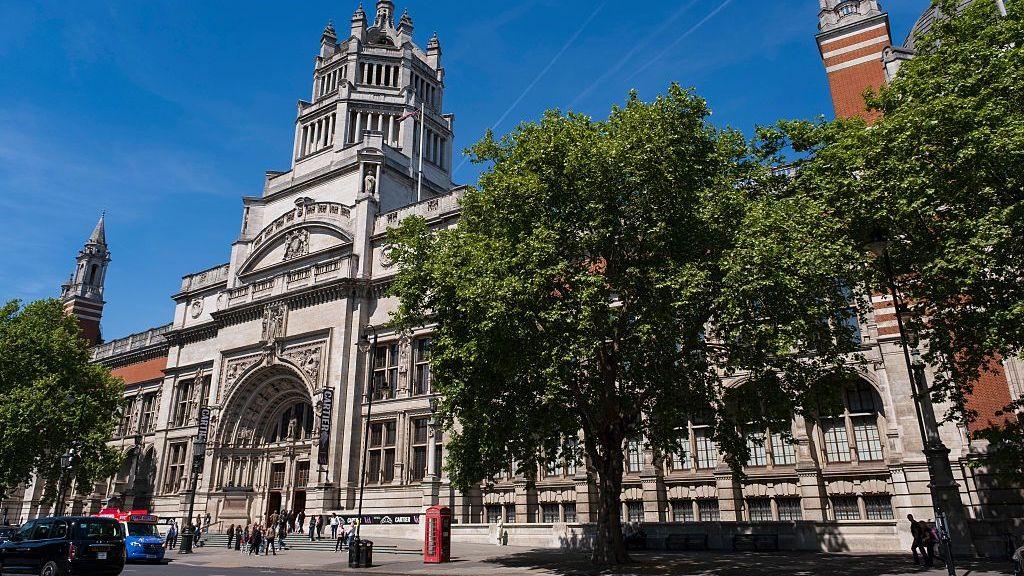 Exterior view of the Victoria and Albert Museum in South Kensington, London, showing its ornate stone entrance, red-brick towers and large trees lining the street on a sunny day.