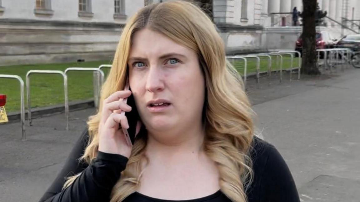 Charlotte Blackwell looks at the camera as she is on the phone. She is standing in front of a court and she has long blonde hair and is wearing a black top.