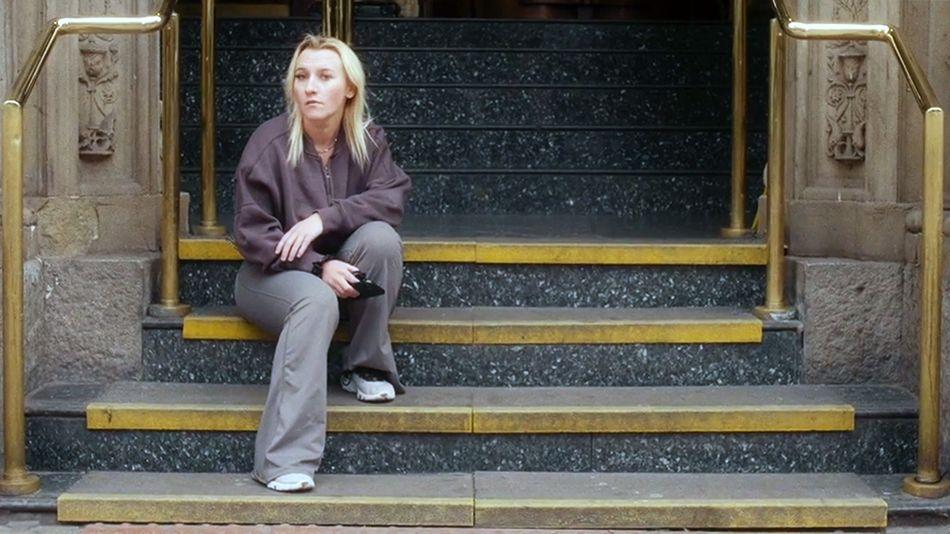 Holly, in casual clothing, sits alone on wide stone steps outside a building entrance. Brass handrails frame the staircase, and dark polished steps lead up to large doors behind her. Holly is holding a phone and looking away from the camera, with the architectural details of the entrance visible in the background.