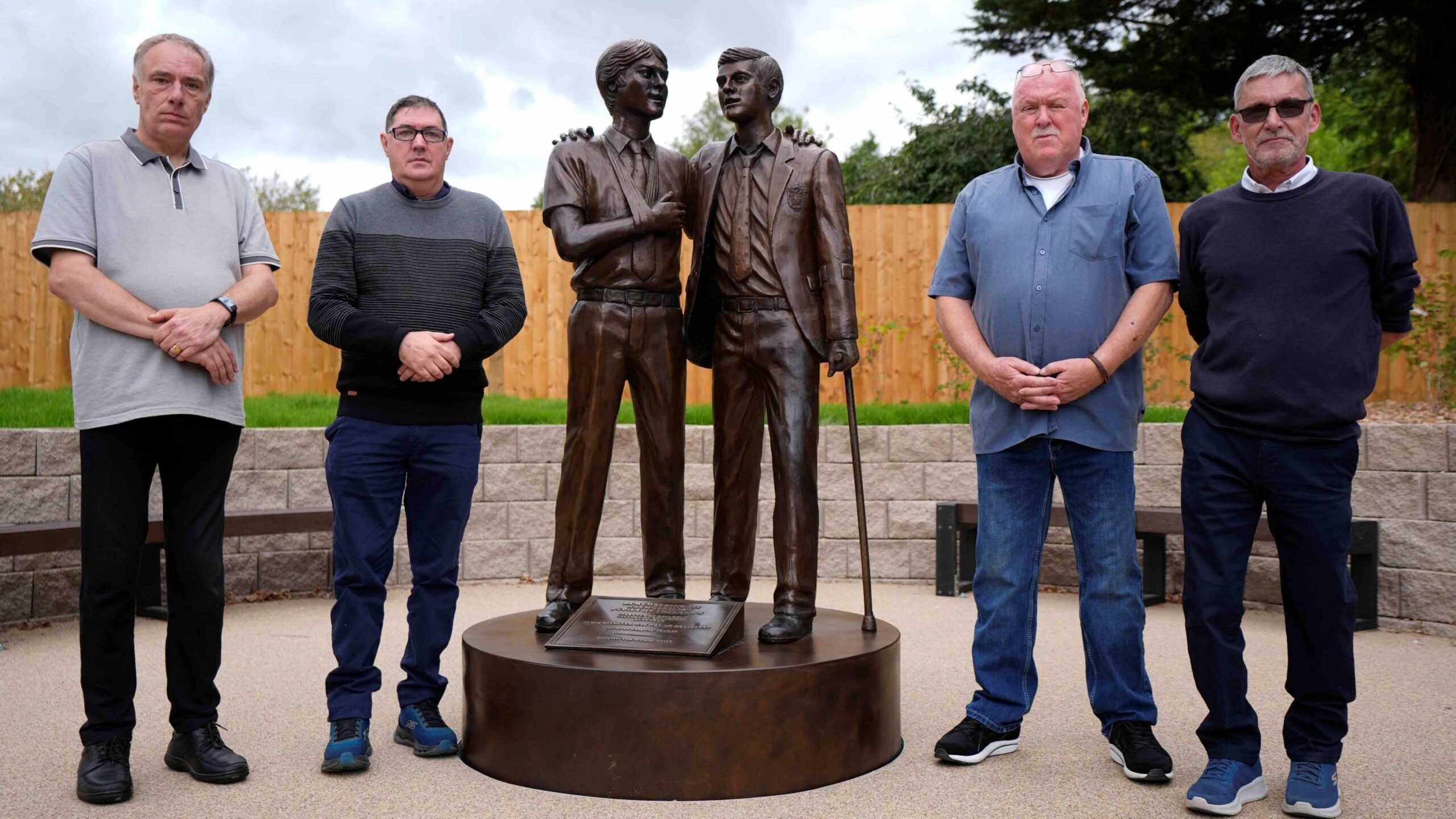 The four men pose next to the bronze memorial, featuring two pupils, one with a sling and and another holding a cane, embracing each other.