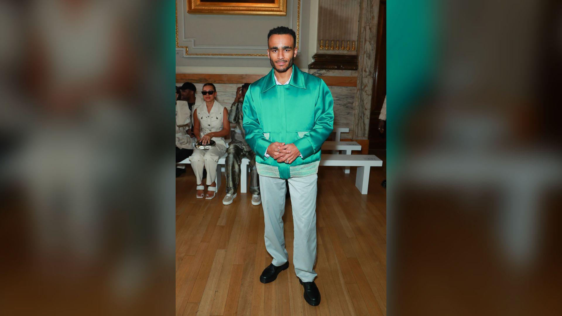 Munya Chawawa has short dark hair hair and is wearing a green zipped-up jacket and light grey trousers.