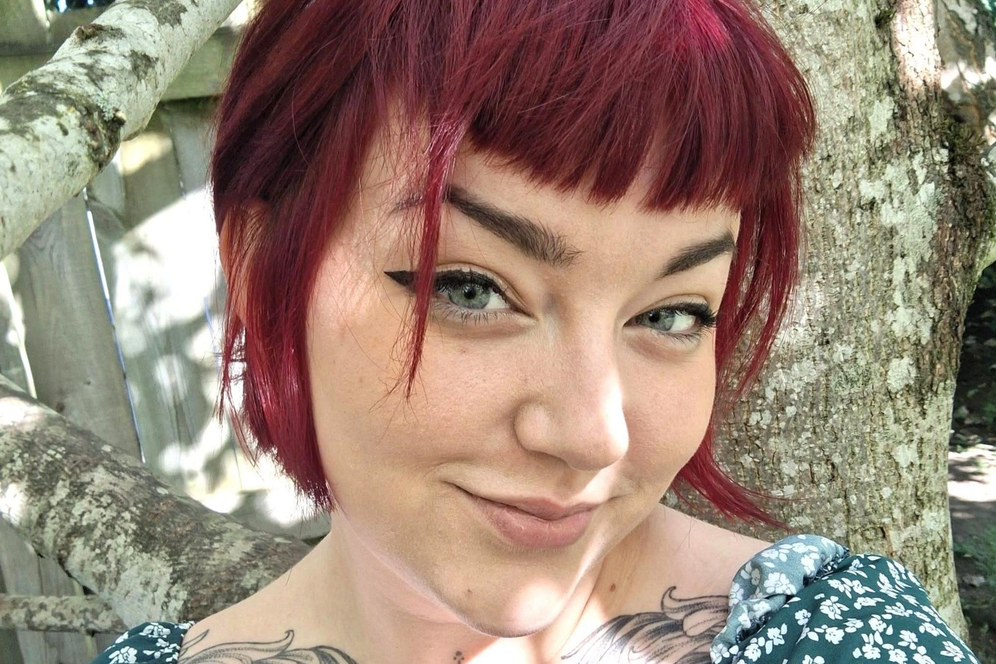 A woman, Charlotte McCann, smiling at the camera, she is wearing a floral top, and she has tattoos visible just below her shoulders.