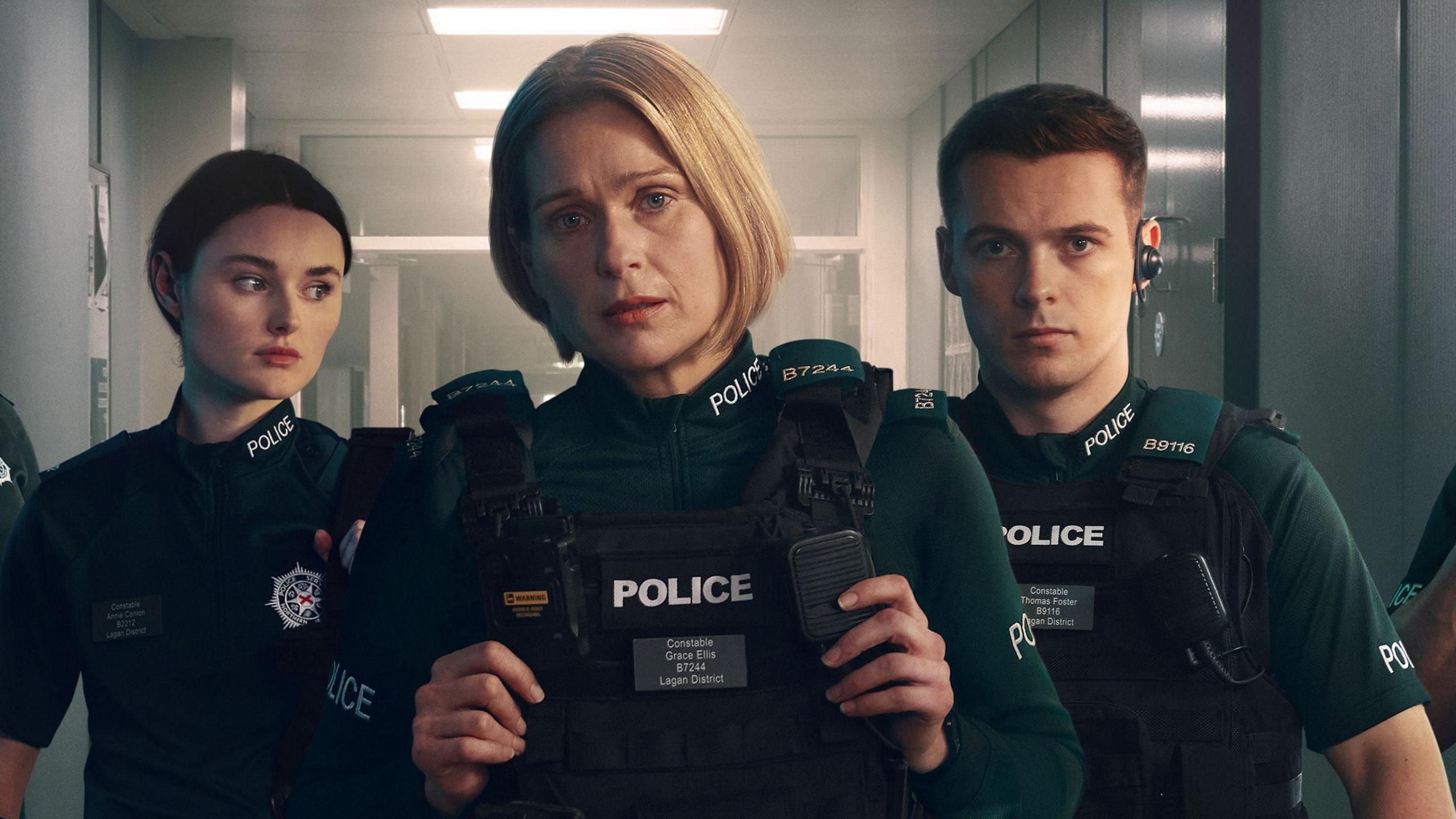 Katherine who plays Annie is standing on the left wearing a dark green police uniform, she has brunette hair tied up and is looking to her left. Siân who plays Grace is standing in the middle and looking straight ahead. She has short blonde hair and is wearing the same uniform. Nathan who plays Tommy is on the right and is also looking straight ahead. He has dark brown short hair. He is wearing the same uniform and has an earpiece in his left ear.