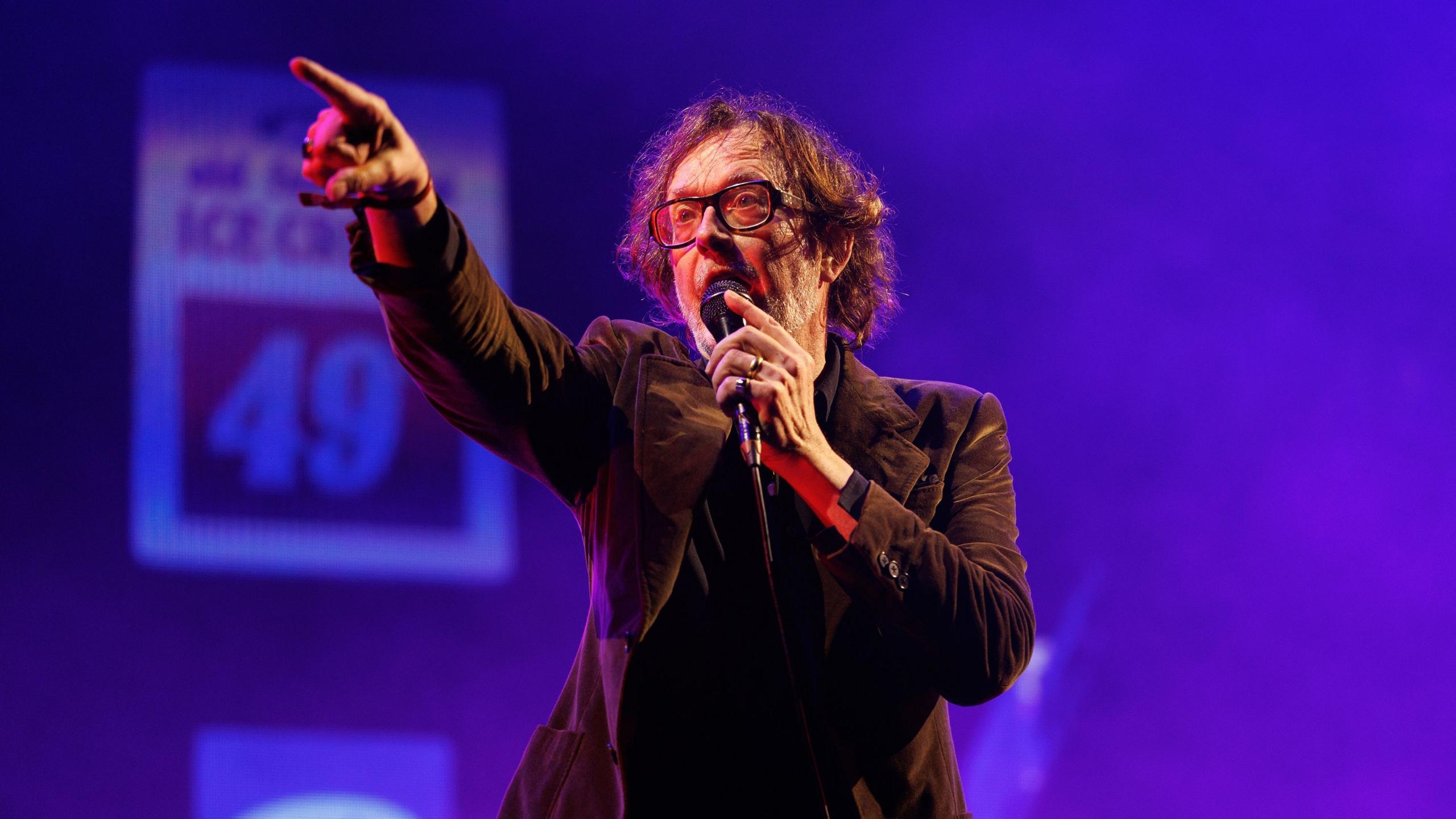 Jarvis Cocker stood on stage singing into a microphone, with his finger points off stage towards the camera and a blue light behind him. He is wearing a brown blazer and glasses.