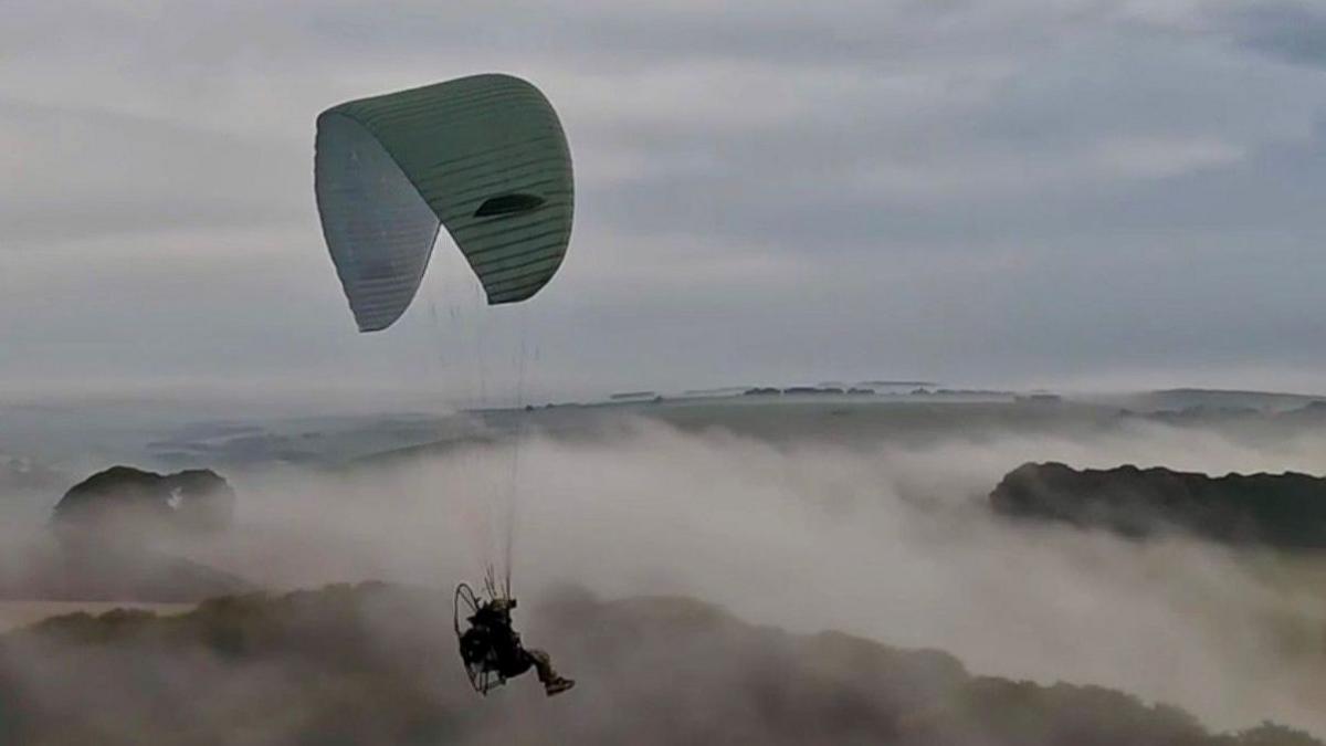 A soldier hanging from a paraglider chute floats through clouds and over a mountain