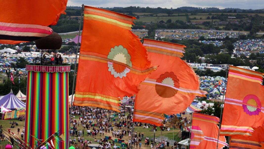 Glastonbury confirms ticket sale dates – and highest ever price Six large orange festival flags fly in the wind. In the background is a tall ribbon tower, which is in one of the field's at Glastonbury festival. Crowds of people can be seen underneath it, with thousands of tents in campsites as far as the eye can see.