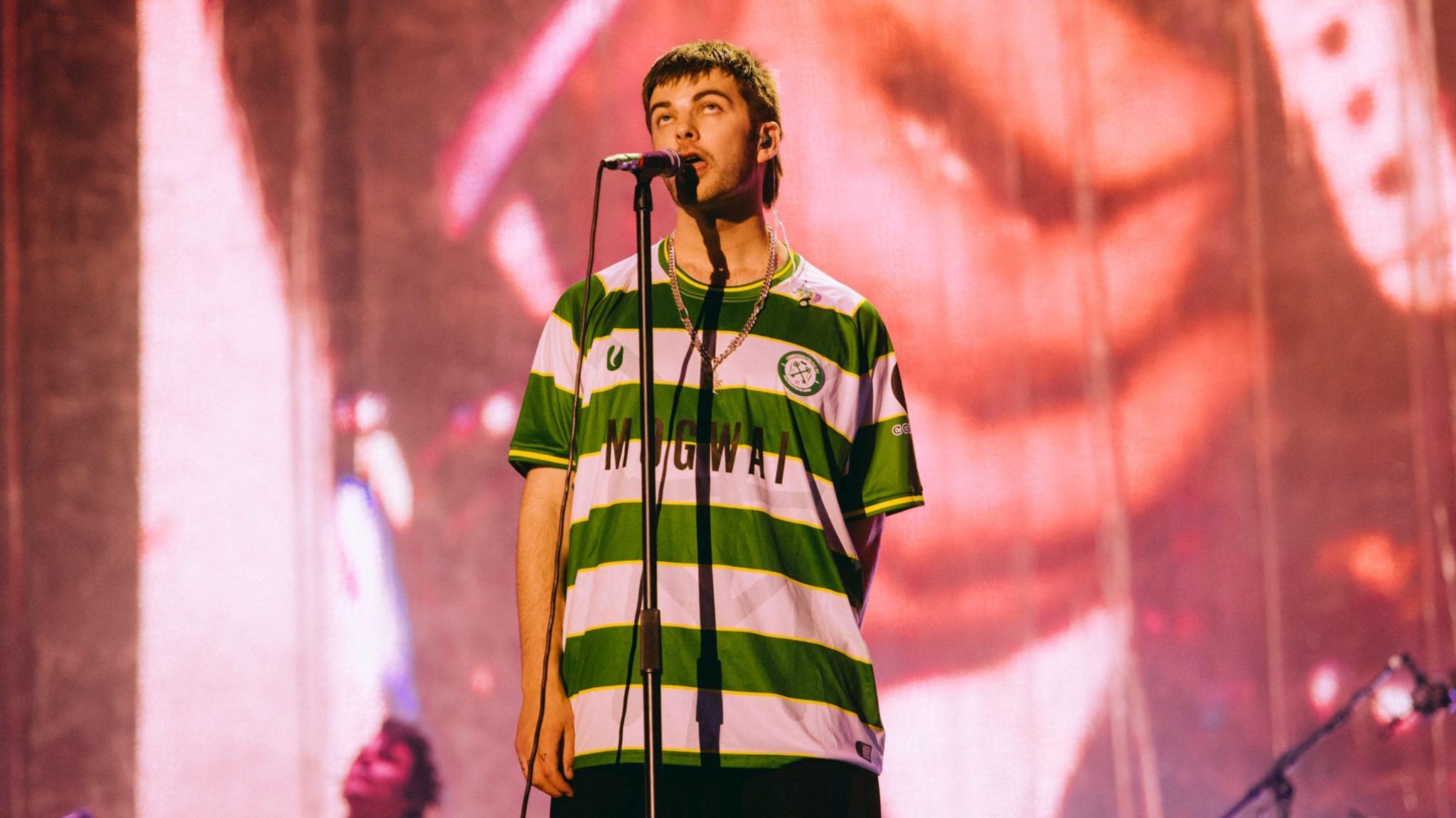 Grian Chatten performing at Reading Festival wearing a Cwmbran Celtic FC away shirt. The football shirt is green and white.