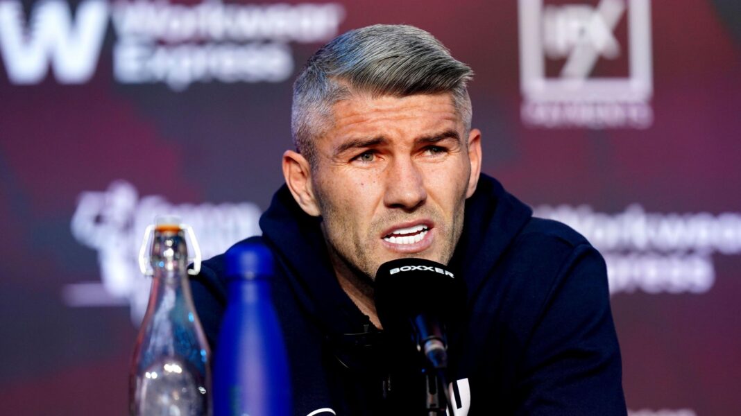 Ex-world champion boxer Liam Smith charged with GBH A man in his 30s, with grey hair and wearing a blue hoody, speaks into a black microphone at a press conference.