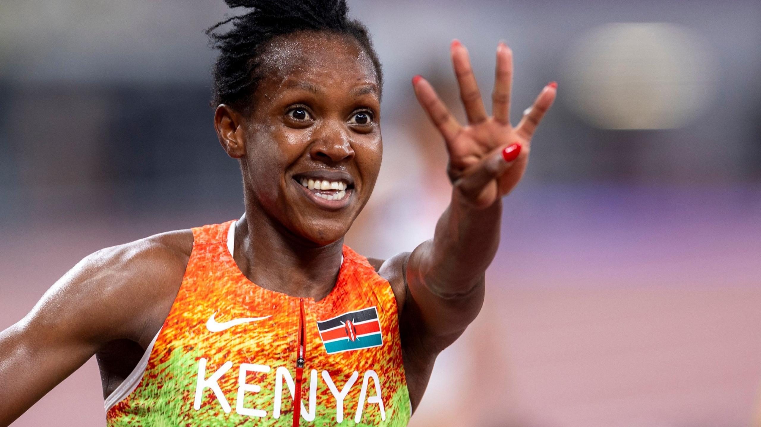 Faith Kipyegon, visible from chest up wearing an orange, yellow and green Kenyan zip-up running top, smiles and holds her left arm in front of her and stretches out four fingers in celebration. The vest has the word 'Kenya' written in white capital text across the chest while a Kenyan flag is on its left breast
