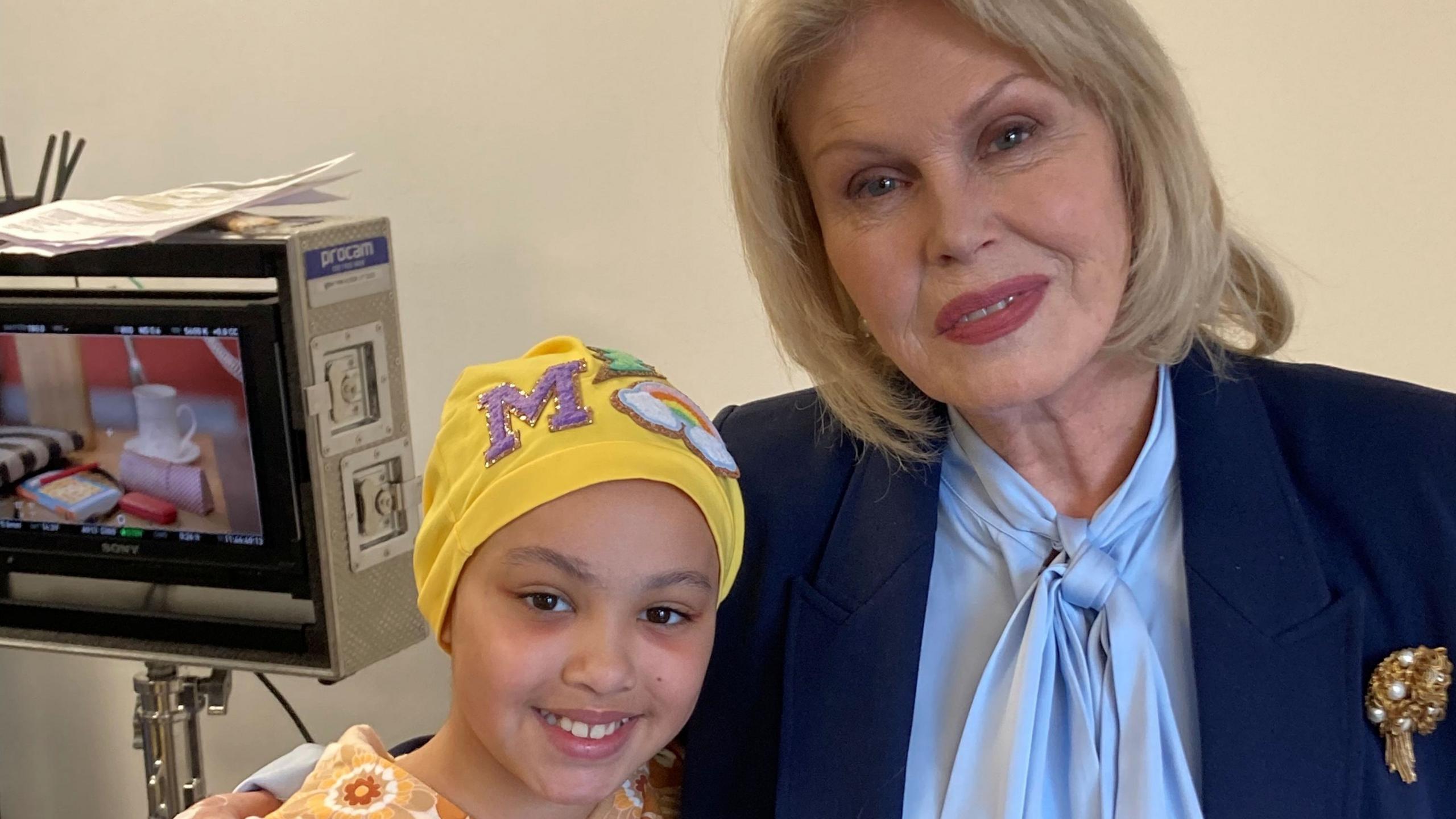 A girl with a yellow head covering with a purple M on the top, and wearing an orange-coloured outfit, smiles as she stands next to Joanna Lumley - a woman with long blonde hair, a blue top and dark blue jacket. A screen can be seen on a stand to the left.