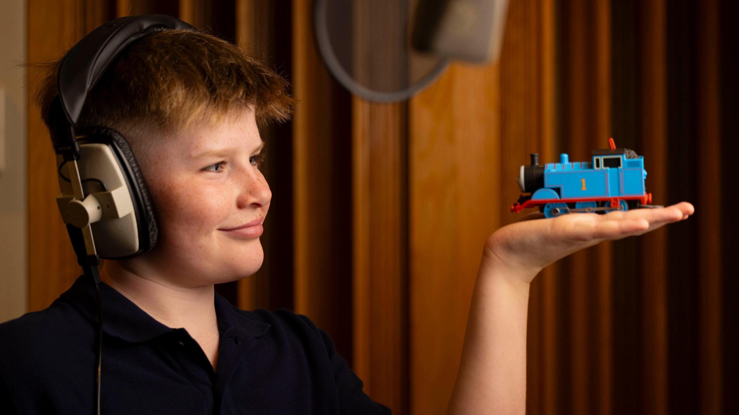 A boy wearing headphones is stood in a recording studio and holding a Thomas the Tank Engine toy on his flat palm.