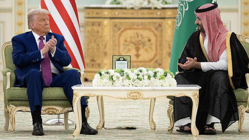 US President Donald Trump and Saudi Crown Prince Mohammed bin Salman sit in green gilded armchairs, the flags of their countries behind them, as they applaud and look at one another. Trump wears a blue suit, white shirt and purple tie while bin Salman wears a black dress and red and white headscarf.